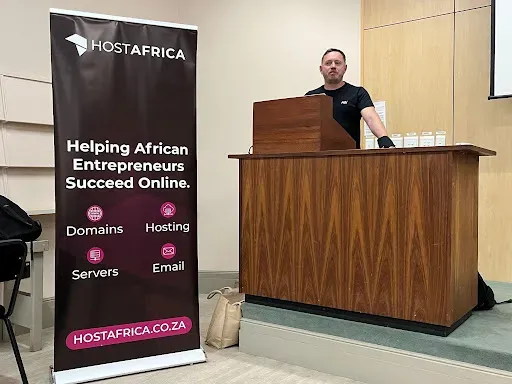 A HostAfrica speaker at the lectern next to a HostAfrica banner listing domains, hosting, servers and email